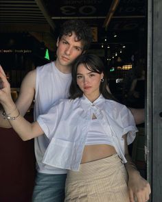 a man standing next to a woman in front of a door wearing a white shirt and tan skirt