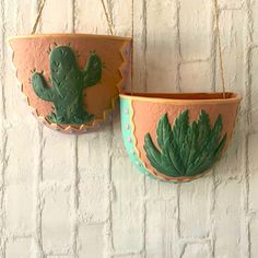 two hanging planters with cactus designs on them against a white brick wall, one is green and the other is pink