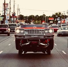 an old car is driving down the street with other cars in the back and on the side