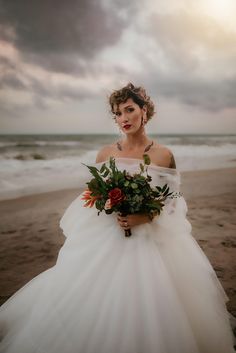 a woman in a white wedding dress holding a bouquet on the beach with dark clouds overhead