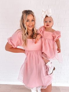 a woman holding a baby wearing a pink dress and white booties, standing in front of a brick wall