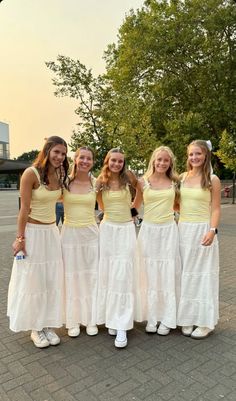 four girls in white and yellow dresses posing for the camera on a brick walkway with trees in the background