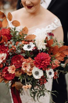 a bride holding a red and white bouquet with greenery in her hand while standing next to the groom