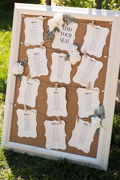 a cork board with seating cards attached to it and flowers hanging from the top, on grass