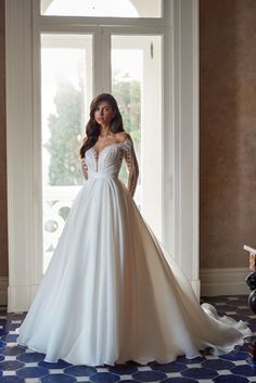 a woman standing in front of a window wearing a white wedding dress with long sleeves