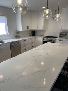 a white kitchen with marble counter tops and stainless steel appliances in the corner, along with hanging lights