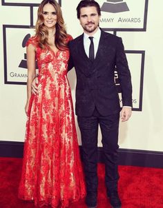 two people standing on a red carpet at an awards event, one in a black suit and the other in a red dress