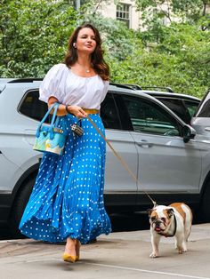 a woman walking her dog on a leash in front of a parked car while wearing a white blouse and blue polka dot skirt