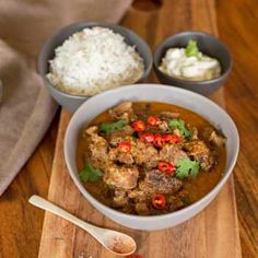 a wooden cutting board topped with a bowl of food and two bowls filled with rice