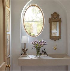 a white sink sitting under a round window next to a table with flowers in it