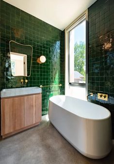 a bath tub sitting next to a window in a green tiled bathroom with wooden cabinets