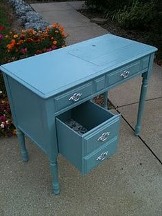 a blue desk with two drawers sitting on the sidewalk next to some flowers and bushes