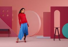 a woman in a red sweater and blue skirt standing next to a pink wall with geometric shapes