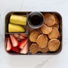 a metal container filled with pancakes, strawberries and bananas next to a cup of coffee