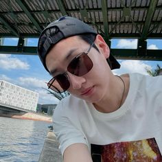 a young man wearing sunglasses and a cap is leaning on a wall near the water