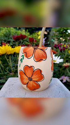 an orange flowered vase sitting on top of a cement slab in front of flowers