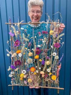 an older woman holding up a piece of art made out of plants and flowers in front of a blue wall