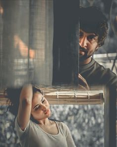 a man and woman standing next to each other in front of a window with sheer curtains