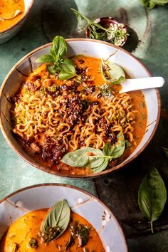 two bowls filled with pasta and basil on top of a green tablecloth next to another bowl full of soup