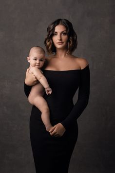 a woman holding a baby in her arms and posing for the camera with an off shoulder black dress