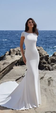 a woman in a white dress standing on rocks near the ocean with her arms behind her back