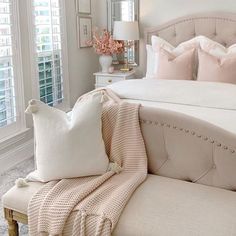 a white bed with pink pillows and blankets on top of it in a bedroom next to two windows