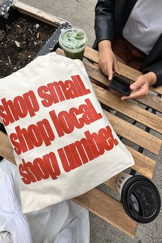 a person sitting on a bench with a tote bag in front of them that says stop small, show local, shop under