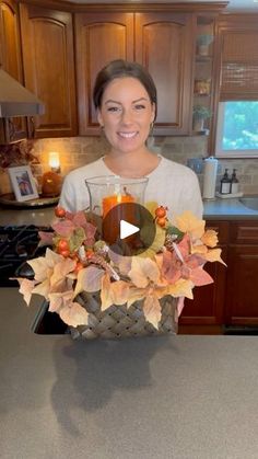 a woman standing in a kitchen holding a basket filled with leaves and an orange candle