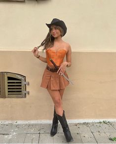 a woman in a short skirt and cowboy hat holding a knife standing next to a wall