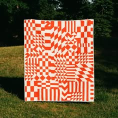 an orange and white quilt sitting on top of a green grass covered field with trees in the background