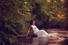 a pregnant woman sitting in the middle of a stream wearing a white dress and holding her belly