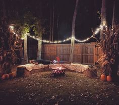 an outdoor fire pit surrounded by pumpkins and trees at night with string lights strung across the fence