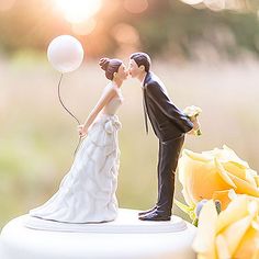 a bride and groom figurine kissing on top of a white cake with yellow flowers