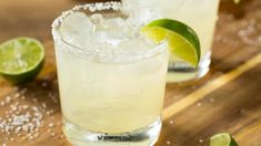 two margaritas sitting on top of a wooden table next to lime slices and salt