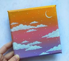 a hand is holding up a small canvas with clouds and the moon painted on it