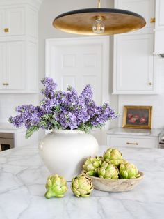 a white vase filled with purple flowers sitting on top of a kitchen counter next to a bowl of artichokes