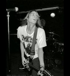 a man with long hair playing guitar in front of a microphone and another person standing behind him