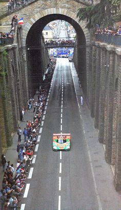 an orange and green car driving down a street with people watching from the sidelines