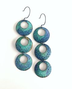 three blue and green circular earrings hanging from hooks on a white background with the word,