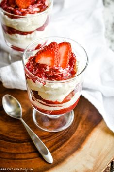 two desserts with strawberries and whipped cream in them on a wooden tray next to spoons