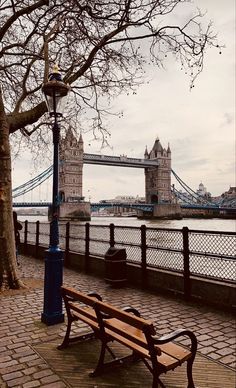 a wooden bench sitting on the side of a river next to a tall tower bridge