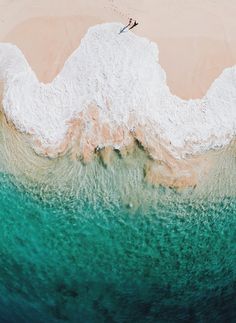 an aerial view of the ocean with two people standing in the water and one person walking on the beach