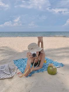 a woman laying on top of a blue and white blanket next to the ocean holding a camera