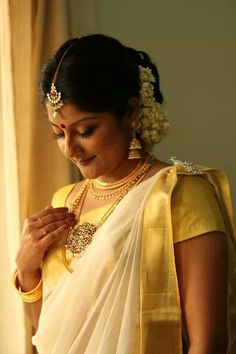 a woman in a yellow and white sari with jewelry on her neck, standing near a window