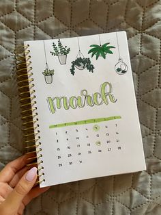 a hand holding a notebook with the word march written in green ink on it and hanging potted plants