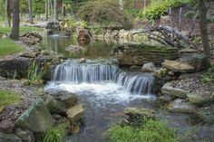 a small waterfall in the middle of a garden with rocks and grass around it, surrounded by trees