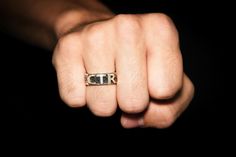 a man's hand wearing a gold ring with the letter t on it, in front of a black background