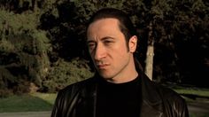 a man in a black shirt and leather jacket looking at the camera with trees in the background