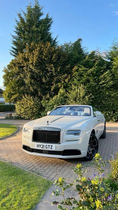 a white rolls royce parked in front of some trees and bushes on a brick walkway