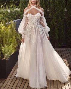 a woman in a white wedding dress standing on a wooden deck with greenery behind her